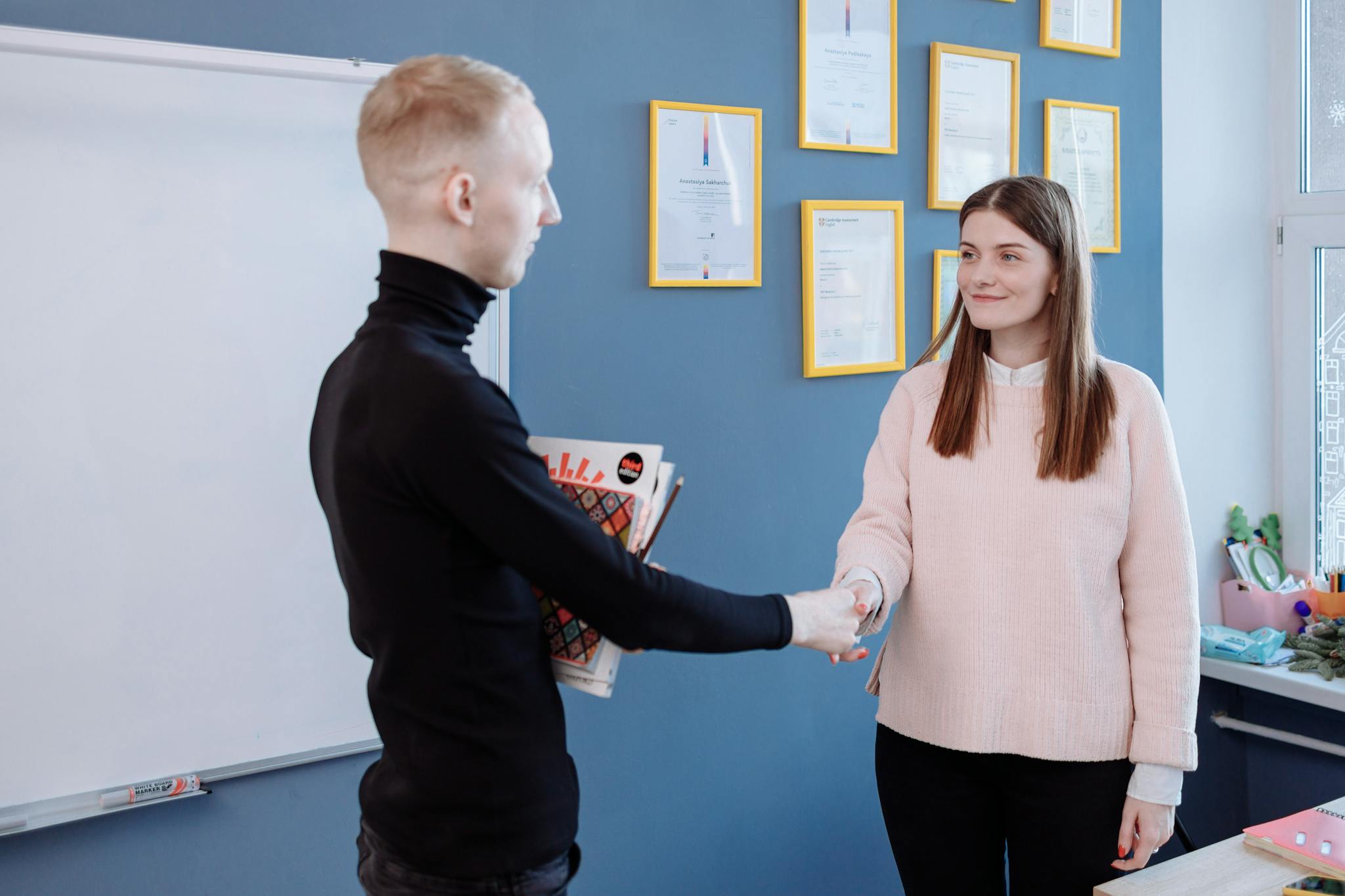Man and woman exchanging handshake in office environment with certificates on wall.