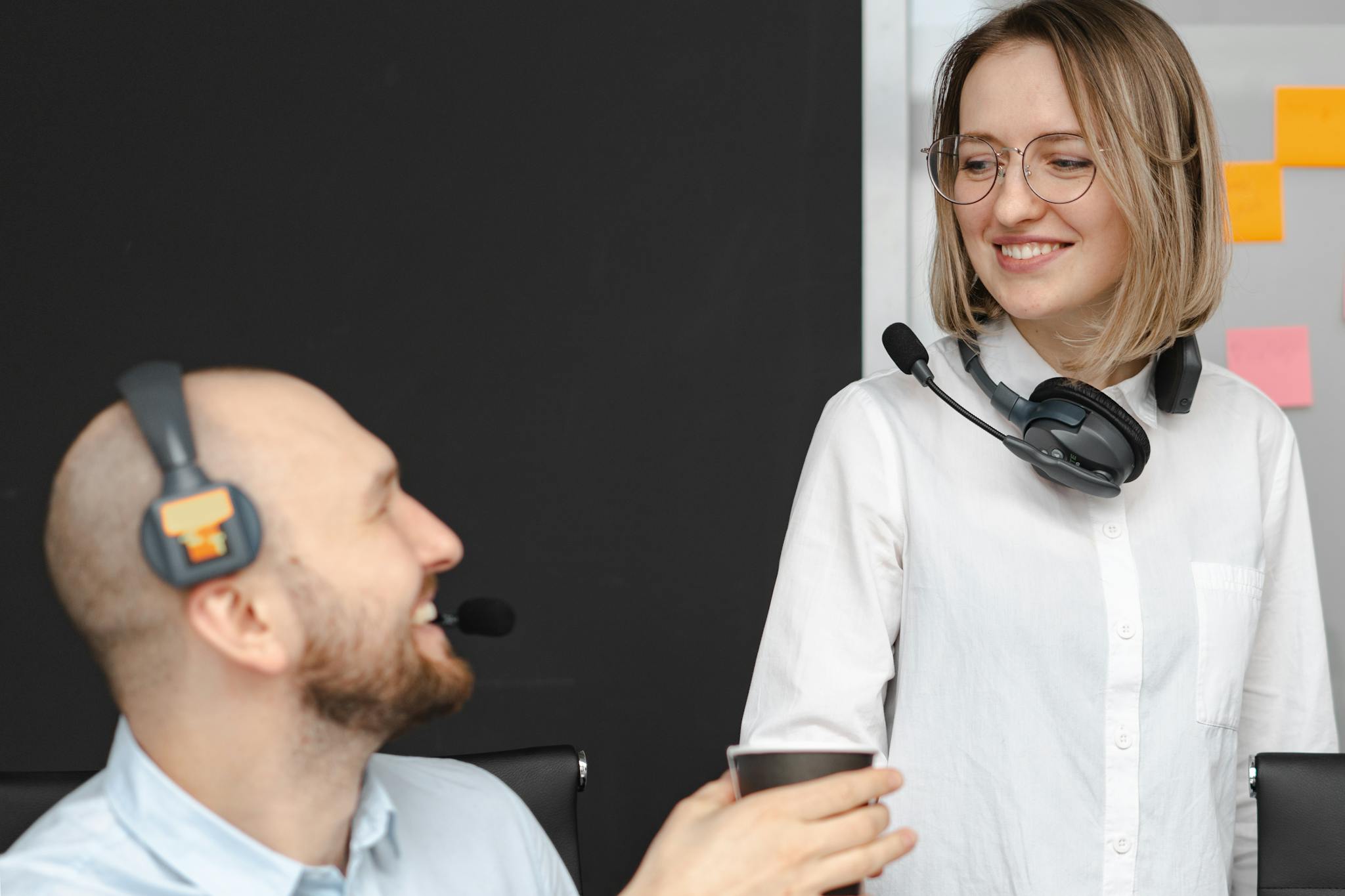 Two call center colleagues with headsets smiling and interacting in an office environment.