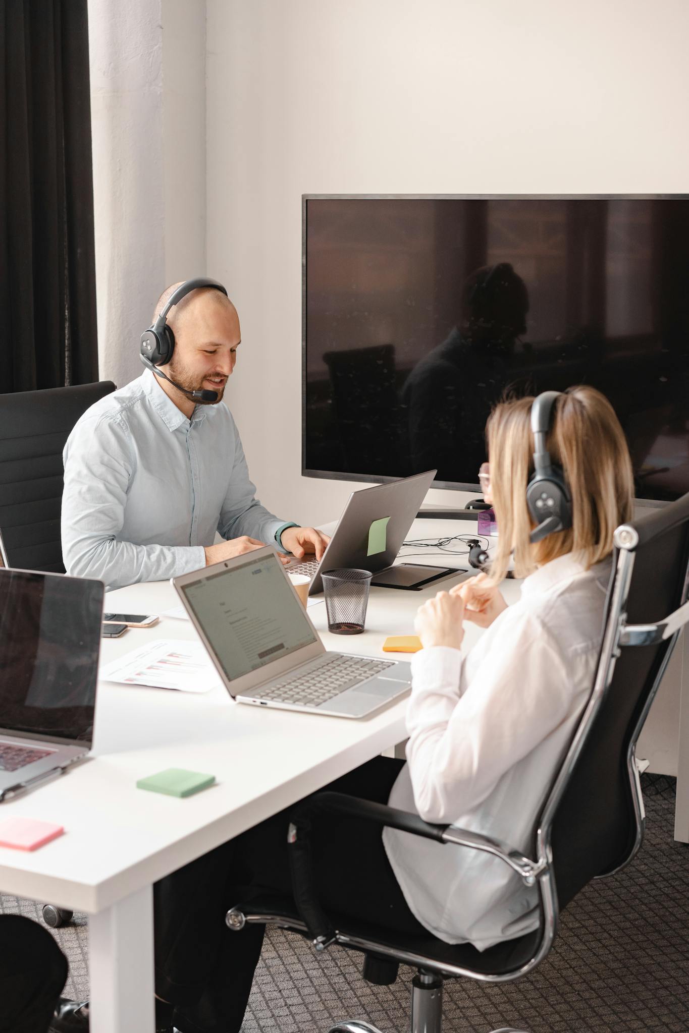 Two office employees in headsets working collaboratively on laptops in a modern workspace.